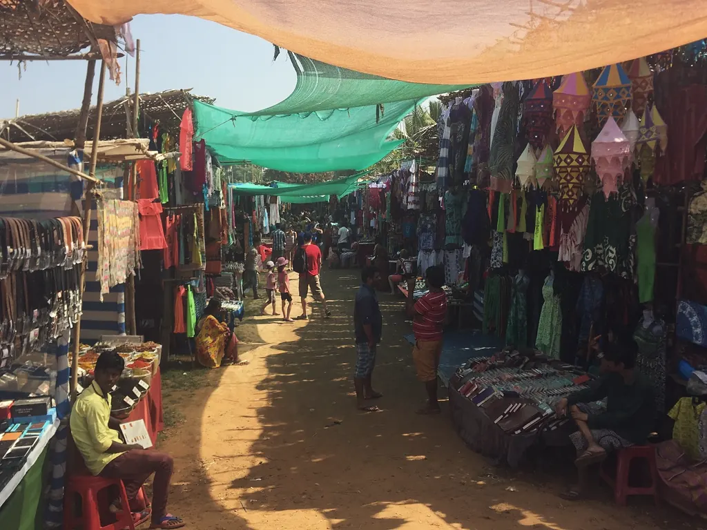 Market stalls with textiles and goods along a shaded lane in Goa